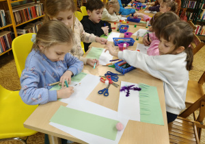 Dzieci siedzą przy stolikach i przy pomocy artykułów plastycznych z zapałem i zaangażowaniem wykonują pracę pt. "Co kryje wiosenna łąka"