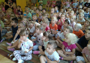 Dzieci z całego przedszkola siedzą na krzesłach i na dywanie i słuchają koncertu