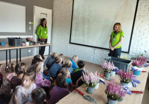 Dzieci siedzą na dywanie i oglądają film edukacyjny o przesadzaniu i pielęgnacji roślin.