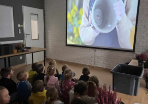 Dzieci siedzą na dywanie i oglądają film edukacyjny o przesadzaniu i pielęgnacji roślin.
