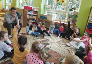 Dzieci wraz z aktorem siedzą w kole na dywanie. Aktor odczytuje zagadki a dzieci chętnie udzielają odpowiedzi