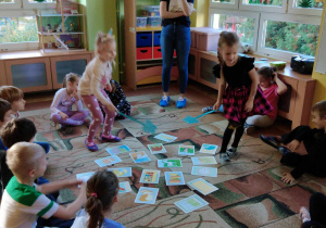 Dzieci siedzą na dywanie, dwie dziewczynki stoją szukają obrazków na określoną głoskę
