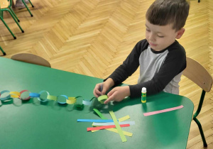 Dzieci siedzą przy stole i kleją choinkowy łańcuch z papierowych kolorowych pasków