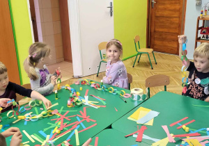 Dzieci siedzą przy stole i kleją choinkowy łańcuch z papierowych kolorowych pasków