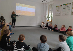 Dzieci siedzą w kole na dywanie, prowadząca omawia prezentacje.