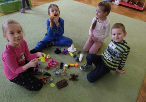 Grupa dzieci siedzi na dywanie i ogląda gotowe przedmioty wydrukowane za pomocą drukarki 3D.