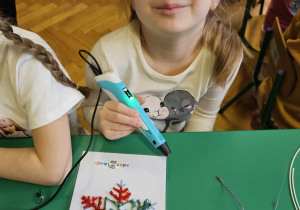 Dzieci podzielone na grupy siedzą przy stolikach i wykonują wzory na szablonach przy użyciu długopisów 3D.