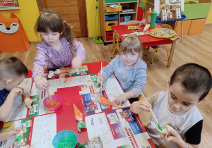 Dzieci siedzą przy stoliku, malują farbami marchewki wykonane z papieru