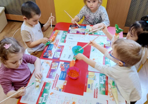 Dzieci siedzą przy stoliku, malują farbami marchewki wykonane z papieru