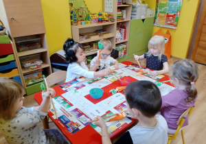 Dzieci siedzą przy stoliku, malują farbami marchewki wykonane z papieru