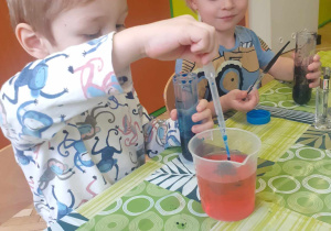 Dzieci siedzą przy stoliku i barwią wodę kolorowym tuszem.