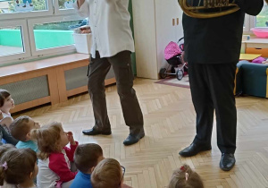 Dzieci siedzą na dywanie, panowie prezentują instrument