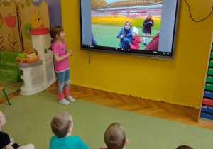 dziewczynka stoi przy tablicy multimedialnej, dzieci siedzą na dywanie i oglądają prezentację