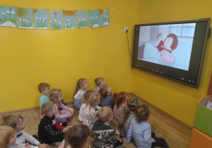 Dzieci z grupy zielonej siedzą przed tablicą multimedialna i oglądają film edukacyjny: "Pierwsza pomoc"