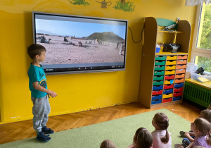 Chłopiec stoi przy tablicy multimedialnej. Dzieci siedzą na dywanie oglądając prezentację