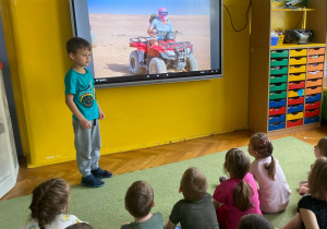 Chłopiec stoi przy tablicy multimedialnej. Dzieci siedzą na dywanie oglądając prezentację