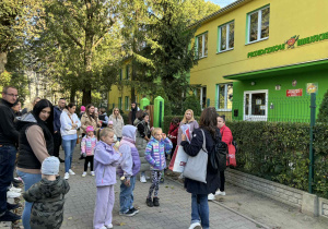 dzieci wraz z rodzicami stoją pod przedszkolem i słuchają od pani przewodnik informacji na temat zadań do wykonania i otrzymują mapki trasy