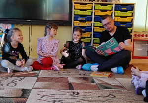 Pan Artur czyta książkę, dzieci siedzą w kole na dywanie słuchając opowieści