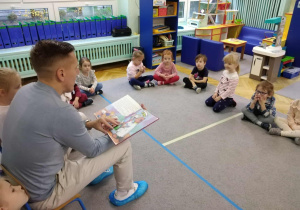 Dzieci siedzą w kole na dywanie, tata Zosi czyta książkę.