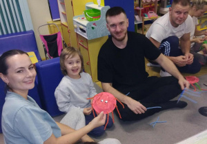 Sara wraz z rodzicami siedzą na dywanie i prezentują wykonanego wspólnie pajączka.