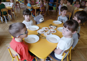 Dzieci wraz z misiem siedzą przy stolikach i jedzą śniadanie