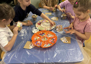 dzieci siedzą przy stołach i ozdabiają tekturowe domki różnorodnym materiałem plastycznym.