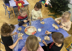 dzieci siedzą przy stołach i ozdabiają tekturowe domki różnorodnym materiałem plastycznym.