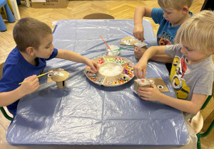 dzieci siedzą przy stołach i ozdabiają tekturowe domki różnorodnym materiałem plastycznym.