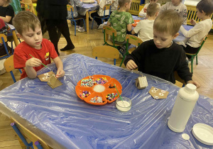 dzieci siedzą przy stołach i ozdabiają tekturowe domki różnorodnym materiałem plastycznym.