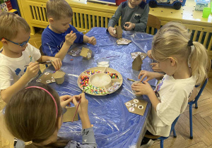 dzieci siedzą przy stołach i ozdabiają tekturowe domki różnorodnym materiałem plastycznym.