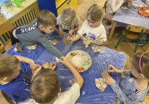 dzieci siedzą przy stołach i ozdabiają tekturowe domki różnorodnym materiałem plastycznym.