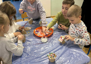 dzieci siedzą przy stołach i ozdabiają tekturowe domki różnorodnym materiałem plastycznym.