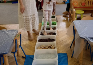 Dziewczynka idzie ścieżką sensoryczną - po gałązkach, szyszkach, kasztanach, kulkach wodnych i wodzie