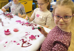 Dzieci siedzą przy stolikach i ozdabiają farbą plakatowa i brokatem serduszka wykonane z masy solnej.