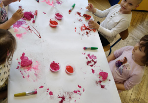 Dzieci siedzą przy stolikach i ozdabiają farbą plakatowa i brokatem serduszka wykonane z masy solnej.