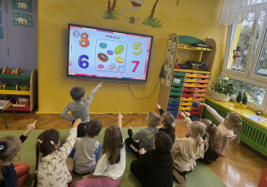 Dzieci siedzą na dywanie i rozwiązują zadania na tablicy multimedialnej