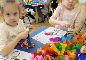 Dzieci siedzą przy stolikach, przy pomocy bibuły i kleju wyklejają baloniki.