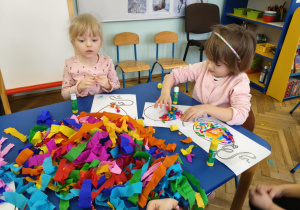 Dzieci siedzą przy stolikach, przy pomocy bibuły i kleju wyklejają baloniki.