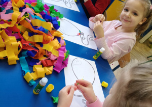 Dzieci siedzą przy stolikach, przy pomocy bibuły i kleju wyklejają baloniki.