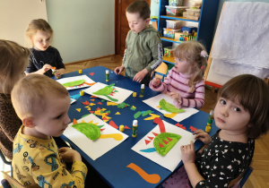 Dzieci siedzą przy stolikach i wykonują pracę plastyczną "Bajkowy dinozaur"