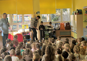 Chłopiec i dziewczynka grają na perkusji, reszta dzieci siedzi w sali i słucha konceru