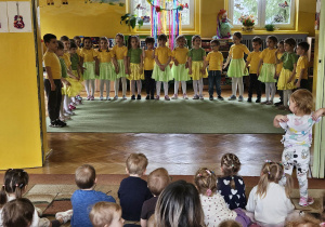 dzieci z grupy żółtej stoją w półkolu i śpiewają piosenkę, na widowni maluchy oglądają występ