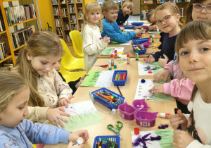 Dzieci siedzą przy stolikach i przy pomocy artykułów plastycznych z zapałem i zaangażowaniem wykonują pracę pt. "Co kryje wiosenna łąka"