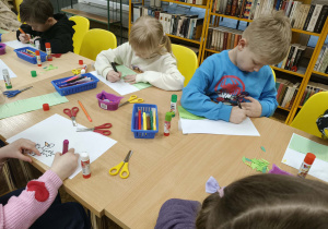 Dzieci siedzą przy stolikach i przy pomocy artykułów plastycznych z zapałem i zaangażowaniem wykonują pracę pt. "Co kryje wiosenna łąka"
