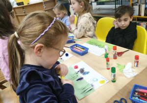 Dzieci siedzą przy stolikach i przy pomocy artykułów plastycznych z zapałem i zaangażowaniem wykonują pracę pt. "Co kryje wiosenna łąka"