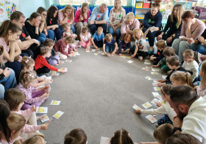 Dzieci wraz z rodzicami siedzą w kole, zabawa "Co było pierwsze, jajko czy kura?"
