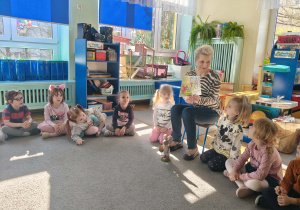 Prowadząca wraz z dziećmi siedzi w kole i czyta dzieciom książkę