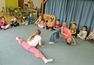 Dzieci siedzą w kole a starsze koleżanki robią ,,mostek" i ,,szpagat"