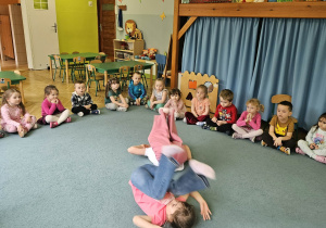 Dzieci siedzą w kole a dwie dziewczynki pokazują elementy akrobatyczne w tym ,,gwiazdę"