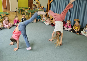 Dzieci siedzą w kole a dwie dziewczynki pokazują elementy akrobatyczne w tym ,,gwiazdę"